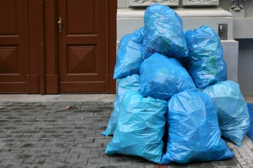 A collection of several large blue plastic rubbish bags, filled with waste, are stacked against a building exterior on a paved driveway. The bags are made of lightweight, semi-transparent plastic, revealing some contents inside, and appear loosely tied at the top. They are arranged in a roughly pyramid shape, with some bags leaning against each other for support. Behind the bags, there is a brown wooden door with a small handle, and part of a white wall with decorative molding is visible in the background. The surrounding ground is paved with interlocking grey bricks, typical of a residential or commercial property exterior. The overall scene depicts a typical rubbish collection described by Commercial Waste Brentwood, serving local needs in the Brentwood area post code, with the bags ready for removal or disposal as part of a waste management service.