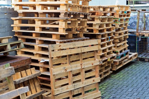 A large stack of wooden pallets arranged neatly outdoors on a paved surface, with some pallets leaning against each other and a few placed on the ground. The pallets are made of light-colored, untreated timber with visible grain and rough textures, and show some signs of wear and use. To the left, there is a partially visible wheelbarrow and additional wooden structures. In the background, a metal fence surrounds the area, which appears to be part of a commercial or industrial yard near Brentwood. The scene is illuminated by natural daylight, highlighting the various materials and textures of the pallets, and emphasizing the context of waste management and rubbish removal services provided by Commercial Waste Brentwood in the local area.