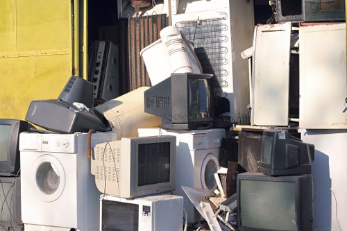 A collection of household appliances and electronic waste, including washing machines, microwave ovens, and small CRT monitors, arranged outdoors against a weathered metal shed or storage unit. The appliances vary in size and colour, predominantly in shades of white, grey, and black, with some showing signs of dirt and age. The washing machines are positioned in the foreground, with some stacked or leaning against each other, while the CRT monitors are scattered nearby, some partially obscured. In the background, there is a rusty metal surface and a yellow panel, indicating an outdoor or industrial environment. The scene is lit with natural daylight, casting soft shadows and highlighting the textures of the appliances, which include smooth plastic, metallic finishes, and glass screens. The image relates to rubbish removal services offered by Commercial Waste Brentwood, focusing on the disposal and clearing of electronic waste and bulky household items from residential or commercial sites.