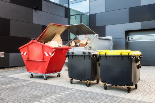 The image shows three wheeled waste bins positioned on a paved surface in an urban setting, with a modern building featuring dark grey paneling and glass windows in the background. The first bin, on the left, is a large red open-top container filled with various cardboard boxes, some of which are partially crushed, revealing its corrugated texture. The middle bin is a dark grey closed container with a yellow lid, situated next to the open red bin, and appears to be made of durable plastic. The third bin, also dark grey with a yellow lid and similar in design, stands further along to the right. Both closed bins are on small castors, facilitating movement, and their surfaces have a matte finish with visible hinges and handles designed for secure closure. The environment suggests a commercial or industrial area, consistent with rubbish removal services available in Brentwood, where Commercial Waste Brentwood operates. The overall scene depicts typical waste management infrastructure, emphasizing the importance of organized rubbish collection for local businesses and properties.